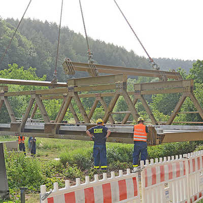 Fachleute des THW nehmen die vormontierte Brücke, die mit einem Kran von der Straße ans Flussufer gehievt wird, „in Empfang“.
