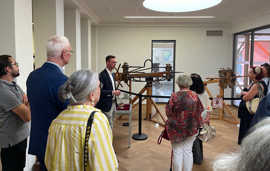 Eine Gruppe schaut sich in einem Museum Militärgeräte aus Holz an. Ein Mann steht in der Mitte und erklärt die Ausstellungsstücke. 