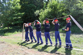 Sechs Jugendliche in Feuerwehrmontur stehen hintereinander auf einer Wiese und halten einen Feuerwehrschlauch über ihre Köpfe.