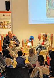 Markus nöhl sitzt in einem Sessel und liest Kindern aus einem Buch vor.
