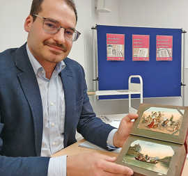 Archivar Jort Blazejewski hat eine gebundene Postkartensammlung aufgeschlagen, zwei kolorierte Bildmotive sind zu sehen.