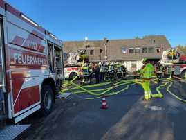 Blick auf den laufenden Feuerwehreinsatz. Am linken Bildrand steht ein Feuerwehrfahrzeug, im Hintergrund das betroffene Haus. In der Bildmitte sind viele Einsatzkräfte.