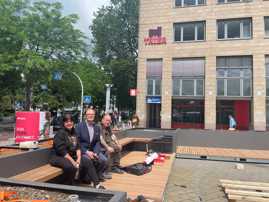 Dezernent Ralf Britten und Projektleiterin Sabine Borkam sitzen gemeinsam mit einem Handwerker auf einer Sitzbank des Parklets. Um sie herum stehen Werkzeuge. Eine Palette liegt vor ihnen auf dem Boden.