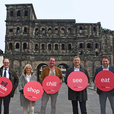 Innenstadt-Dezernent Ralf Britten, Karin Kaltenkirchen, Vertreterin für den Einzelhandel,  Patrick Sterzenbach, Vorsitzender der City-Initiative Trier, TTM-Chef Norbert Käthler und Alexander Brittnacher, Vertreter der Gastronomie und Vorstandsmitglied der City-Initiative, stehen vor der Porta Nigra. Sie halten große, runde Schilder hoch, auf denen die Schlagwörter der neuen Marketingkampagne „Shop“, „Chill“, „See“ und „Eat“ stehen.