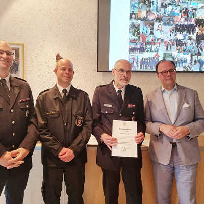 Andreas Resch (2. v. r.) präsentiert die Urkunde zum Abschied aus dem aktiven Dienst, die ihm Dezernent Ralf Britten, Dr. Andreas Palzer, Vize-Chef der Berufsfeuerwehr, und Stephan Kuhn, Leiter der Freiwilligen Feuerwehr Kürenz, (v. r.) übergeben haben. Die Präsentation im Hintergrund zeigt neben vielen persönlichen Erinnerungen von Andreas Resch auch, wie stark sich die Ausstattung in den letzten Jahrzehnten gewandelt hat. 