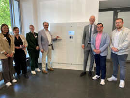 Sieben Personen, darunter Oberbürgermeister Wolfram Leibe und Dezernent Ralf Britten stehen vor einer circa 1,80 Meter hohen Box, die insgesamt 304 kleine Fächer aufweist. Eines davon ist auf und Dezernent Britten tut, als ob er seinen Ausweis aus diesem Fach nimmt.  