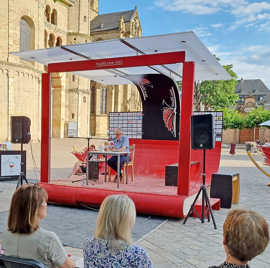 Eine Lesung findet auf einer roten Bühne statt, die auf einem Platz vor einer Kirche steht.