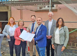 Vor dem vor rund 100 Jahren errichteten Gebäude Magnerichstraße 9-11 überreicht Doris Ahnen den Förderbescheid an Steve Klein und Stefan Blankenberg. Mit dabei sind außerdem Sandra Baumbach, Wolfram Leibe und Sybille Jeschonek 