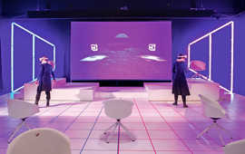 Blick in einen virtuellen Bühnenraum mit zwei Figuren, Sitzmöbeln vor einem Hintergrund in violetten Farbtönen