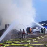 Auf dem Bild sieht man Wasser, das aus mehreren Schläuchen auf einen großen, qualmenden Haufen gespritzt wird. Schläuche sind am Boden verteilt. Feuerwehrleute im Einsatz.