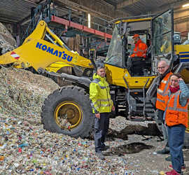 Ein Arbeiter im Radlader bewegt Berge von Plastikmüll und vor dem Fahrzreug stehen drei Besucher mit Warnwesten