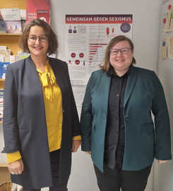 Angelika Winter und Hannah Grunewald stehen in einem Büro vor einem Plakat mit der Aufschrift "Gemeinsam gergen Sexismus"