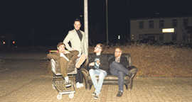 Drei Mitglieder der Band „Papertones“ sitzen nachts unter einer Straßenlaterne auf einem Sofa, ein weiterer liegt in einem Einkaufswagen.