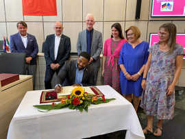 Ein Mann trägt sich handschriftlich in ein Buch ein, das auf einem Tisch mit Blumenschmuck liegt. Im Hintergurnd stehen weitere Personen.