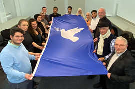 Die Teilnehmerinnen und Teilnehmer des Dialogs im Rathaus halten gereinsam eine blaue Fahne mit einer weißen Friedenstaube