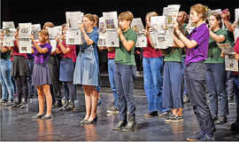 Auf einer Bühne steht eine große Gruppe Jugendlicher. Sie sind alle in dieselbe Richtung gewandt und halten aufgeschlagene Zeitungen vor ihren Gesichtern.