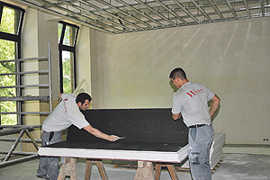 Zwei Mitarbeiter einer Spezialfirma schneiden Schallschutzplatten zu, die an einer Metallkonstruktion an der Decke befestigt werden.