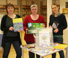 Die APAG-Projektmitarbeiterinnen Ruth Strauß (links) und Natalie Smilek (rechts) übergeben ihrer Bibliothekskollegin Iris Gesellchen die Materialkiste, die für die Lernpaten bereit steht. Foto: Stadtbibliothek