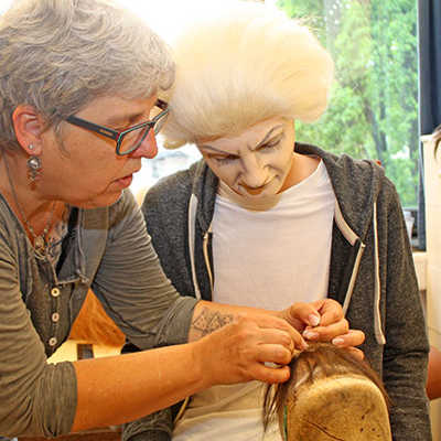 Maskenbildnerin Birgit Molitor zeigt dem bereits geschminkten Ben, wie im Theater Haarteile geknüpft werden.