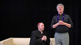 Ein als Kardinal verkleideter Schauspieler kniet neben einem ebenfalls als Geistlicher verkleideten Schauspieler, der mit geschlossenen Augen und gefalteten Händen dasteht.