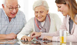 Zwei Senioren spielen mit einer jüngeren Dame Puzzle und lächeln.