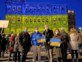 Die Dezernenten Markus Nöhl und Ralph Britten stehen gemeinsam mit Oberbürgermeister Wolfram Leibe, Bürgermeisterin Elvira Garbes und vielen weiteren Bürgerinnen und Bürgern vor der blau-gelb beleuchteten Porta Nigra.