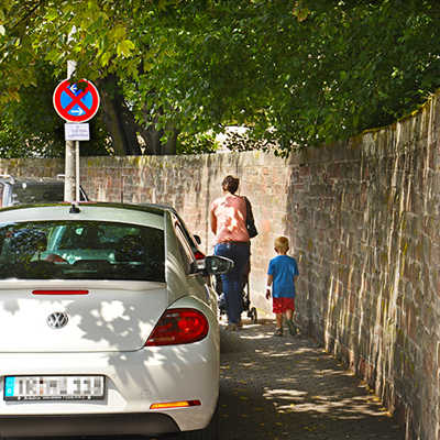 In der Mustorstraße parkt ein Autro trotz Verbot auf dem Fußweg, sodass eine Mutter mit Kinderwagen und ihrem älteren Sohn kaum noch vorbeikommt.