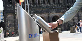 Aus einem Wasserspender wird eine Trinkfalsche befüllt vor dem Hintergrund eines römischen Bauwerks