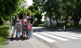 Vier Erewachsene und drei Kinder stehen auf einem Gehsteig, von dem aus ein Zebrastreifen über die Straße führt.