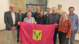 Gruppenfotot im Rathaus-Foyer, die Personen halten eine große rote Flagge mit Trauben-Symbolen in die Höhe