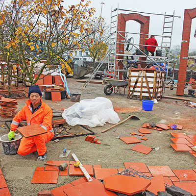 Ein Gartenarbeiter aus Xiamen hält eine der aus China importierten Ziegelplatten, die auf der Plattform des Partnerschaftsgartens verlegt werden. Im Hintergrund wird an einem der Windschirme gearbeitet.