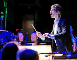 Ein Dirigent steht vor einem Orchester. In seiner Hand hält er einen Taktstock und schaut konzentriert noch vorne. Er wird von unten angeleuchtet. Im Hintergrund sieht man einige Musiker.