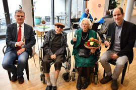 Geburtstagsfeier für Anneliese Mettlach mit Gratulanten, Blumenstrauß und Sekt. Sie hat weiße Haare, trägt ein grünes Kostüm und sitzt in einem Rollstuhl.