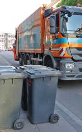 Ein Müllauto hält neben mehreren Restmülltonnen. Im Hintergrund steht die Porta Nigra.