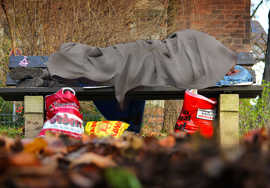 Ein Mensch liegt in eine Decke gehüllt auf einer Parkbank, vor der mehrere Plastiktüten stehen.
