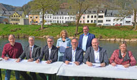 Sieben Vertreterinnen und Vertreter der beteiligten Kommen und Verbände sitzen nebeneinander an Tischen auf einer Wiese am Flussufer und unterzeichnen Dokumente.