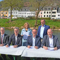 Sieben Vertreterinnen und Vertreter der beteiligten Kommen und Verbände sitzen nebeneinander an Tischen auf einer Wiese am Flussufer und unterzeichnen Dokumente.