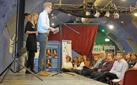 Drei Personen stehen auf einer Bühne, einer von ihnen spricht in ein vor ihm aufgestelltes Mikro. Vor der Bühne sitzen mehrere Menschen und blicken interessiert zu dem Redner herauf.