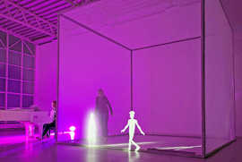 Szene aus dem Stück: Der Raum ist mit lila Licht beleuchtet, in der Mitte steht ein vier mal vier mal Meter großen Kubus, der als Projektionsfläche dient. 