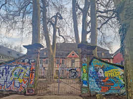 Mauereinlass zum mit Bäumen bestandenen Hof eines historischen Gebäudes, zu beiden Seiten mit Graffiti bemalt.