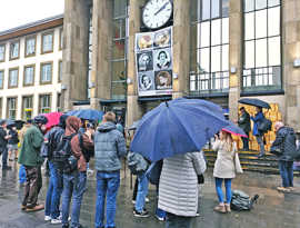 Besucherinnen und Besucher der Gedenkveranstaltung stehen vor dem Ausstellungsplakat am Eingang des Hauptbahnhofs