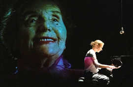Ein Frau spielt in einem abgedunkelten Raum Klavier vor dem Hintergrund der Video-Projektion des Gesichts einer älteren Frau.