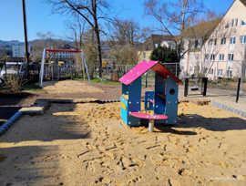 Zu sehen ist ein Spielplatz mit einem Spielhaus, das im Sandkasten steht. Im Hintergrund ist eine Schaukel zu sehen.