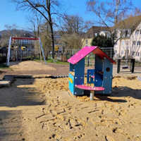 Zu sehen ist ein Spielplatz mit einem Spielhaus, das im Sandkasten steht. Im Hintergrund ist eine Schaukel zu sehen.