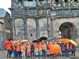 Eine Gruppe Frauen, die orangefarbene Kleidungsstücke und Regenschirme tragen, steht vor der Porta Nigra, an der ein Plakat angebracht wurde