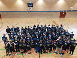 Die Teilnehmerinnen und Teilnehmer des Völkerballturniuers stehen in eier Turnhalle und tragen einheiltiche dunkelblaue T-Shirts.mit Aufdruck auf dem Rücken