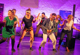 Das Bild zeigt eine Tanzgruppe auf einer in violettes Licht getauchten Bühne