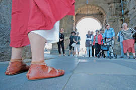 Das Bild zeigt im Anschnitt eine Person in altrömischer Kleidung und im hintergrund die Teilnehmer einer Stadtführung