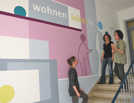 Jean Martin Solt (Verein Transcultur), Annika Rieche (Club Aktiv) und die Designerin Gabi Bruckmann  (v. r.) präsentieren eine neugestaltete Wand in der Abteilung für Wohnungswesen des Sozialamtes.