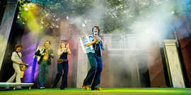 Theaterszenenfoto mit vier Schauspielern, die als Musikgruppe mit verschiedenen Instrumenten und Kostümen im Stil der 70er Jahre auftreten.